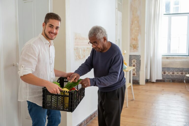 A young volunteer offers support by delivering fresh vegetables to an elderly gentleman indoors.