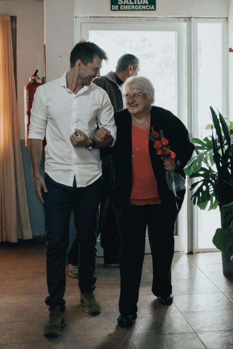 Senior woman arm in arm with a man, smiling and walking indoors with a bouquet of flowers.
