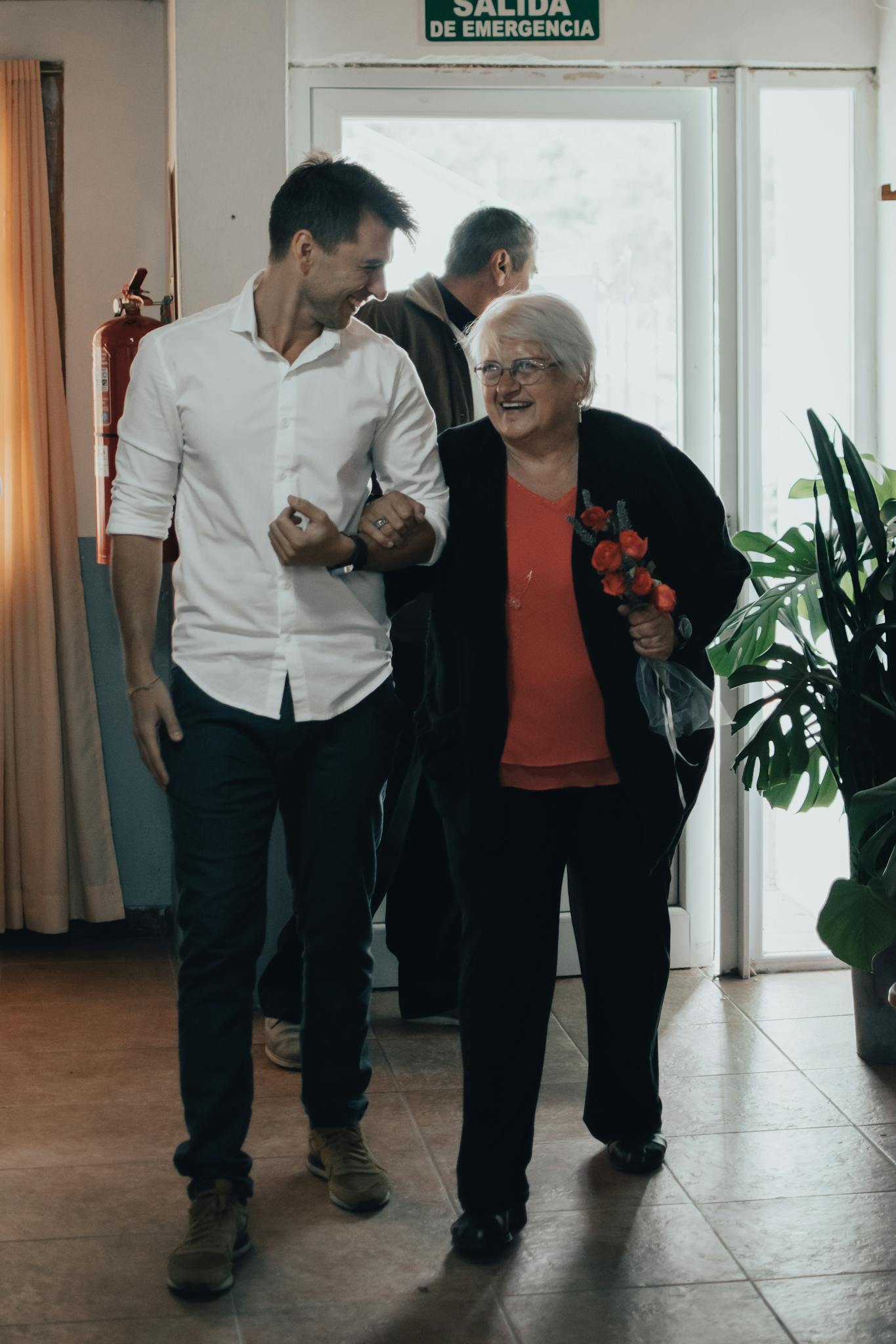Senior woman arm in arm with a man, smiling and walking indoors with a bouquet of flowers.